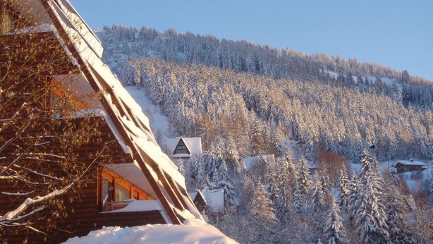 Una cabaña de madera con techo inclinado, rodeada de árboles cubiertos de nieve y un cielo azul despejado.