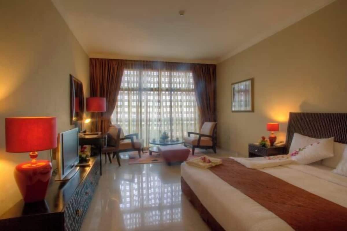 A hotel room with a bed, a desk with a computer, a chair, a small table, and a window with curtains.