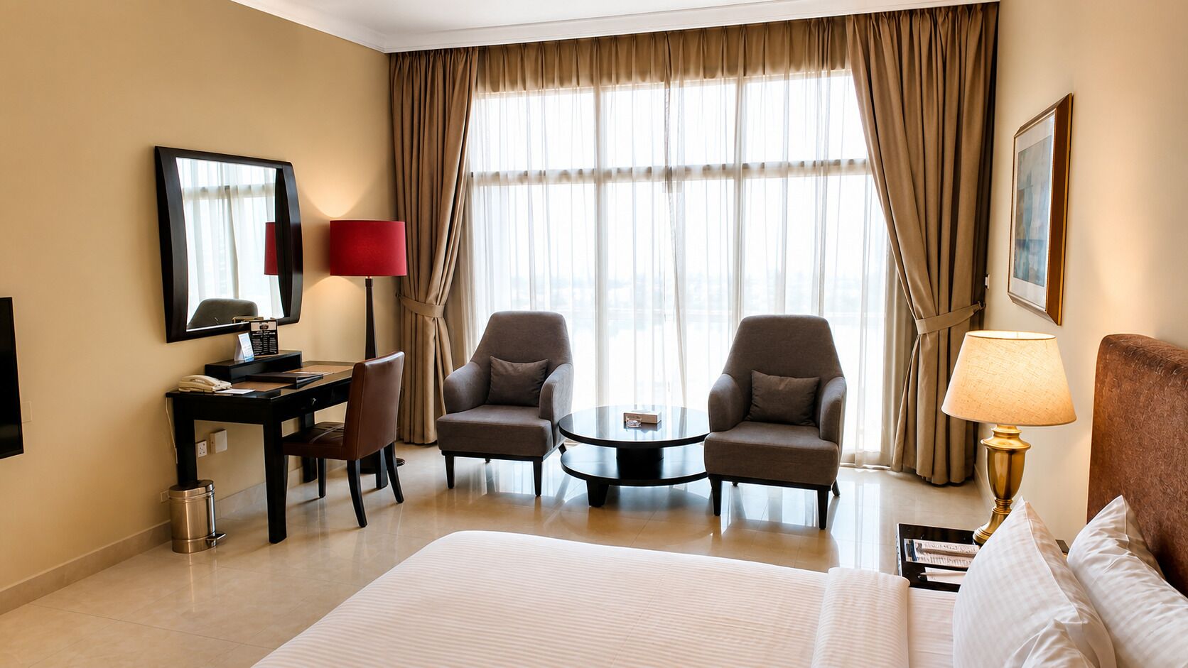 A hotel room with a large bed, two armchairs, a small table, a desk with a lamp, and a large window with curtains.