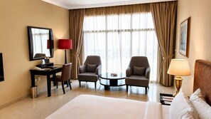 A hotel room with a large bed, two armchairs, a small table, a desk with a lamp, and a large window with curtains. - Oryx Hotel (Abu Dhabi)