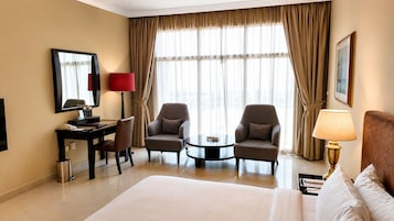Habitación de hotel con una cama grande, dos sillones, una mesita, un escritorio con lámpara y un ventanal con cortinas.