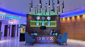 A hotel reception desk with a digital screen, two reception chairs, and a wall with clocks and framed pictures. - Oryx Hotel (Abu Dhabi)