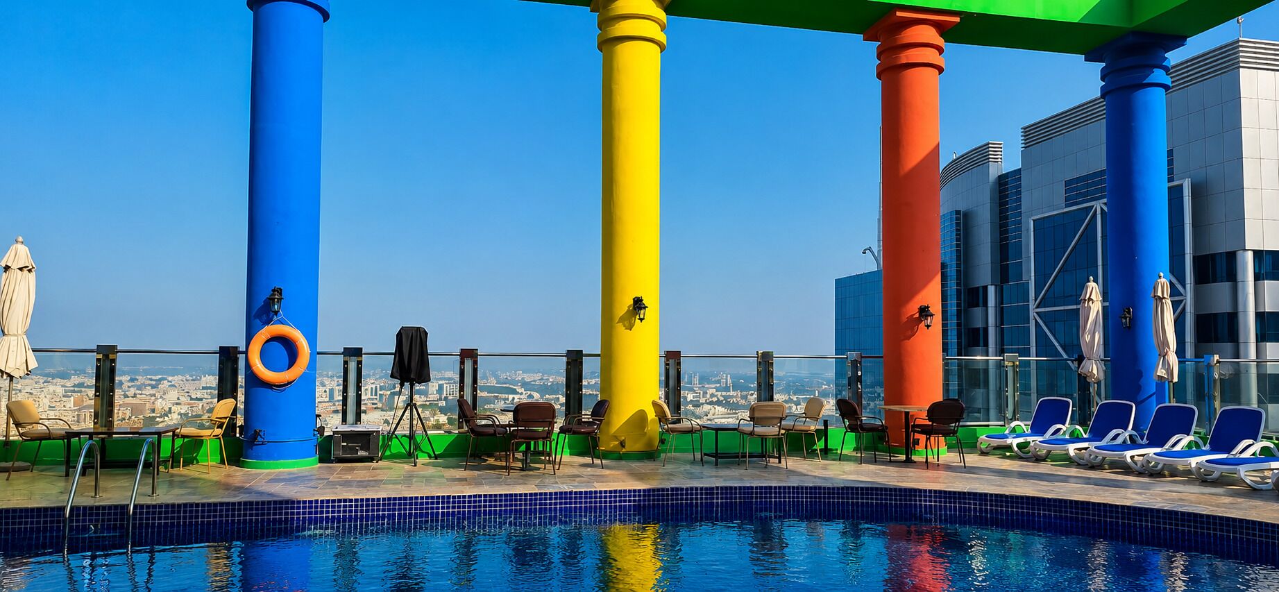 A rooftop pool area with a clear view of the cityscape, featuring a pool, lounge chairs, and a railing.