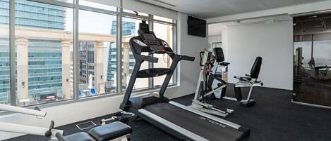 Un gimnasio moderno con cintas de correr, una banca y equipo de ejercicio. Amplias ventanas ofrecen vistas a un paisaje urbano.