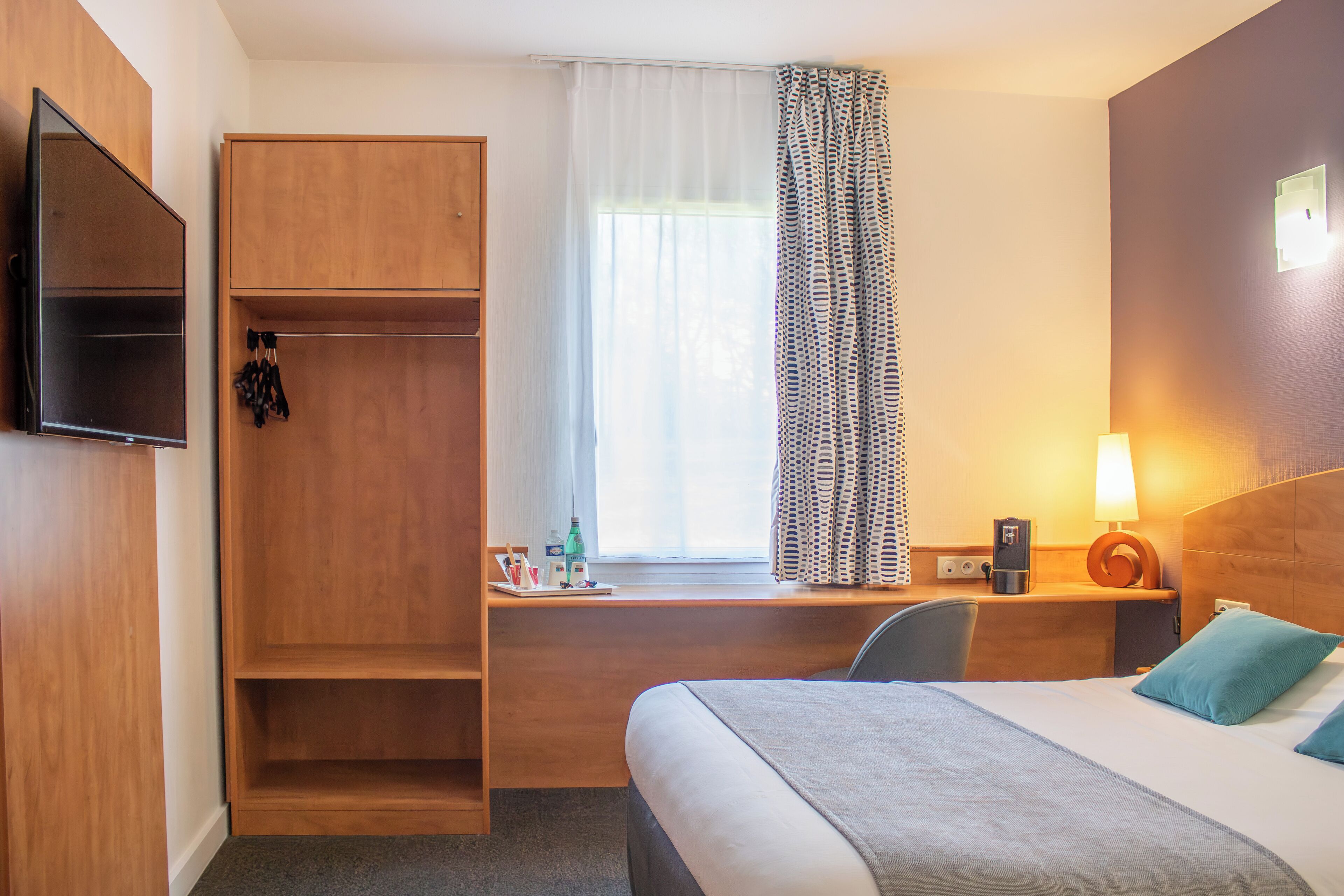 A hotel room with a bed, a desk with a chair, a wardrobe, a television, and a window with curtains.