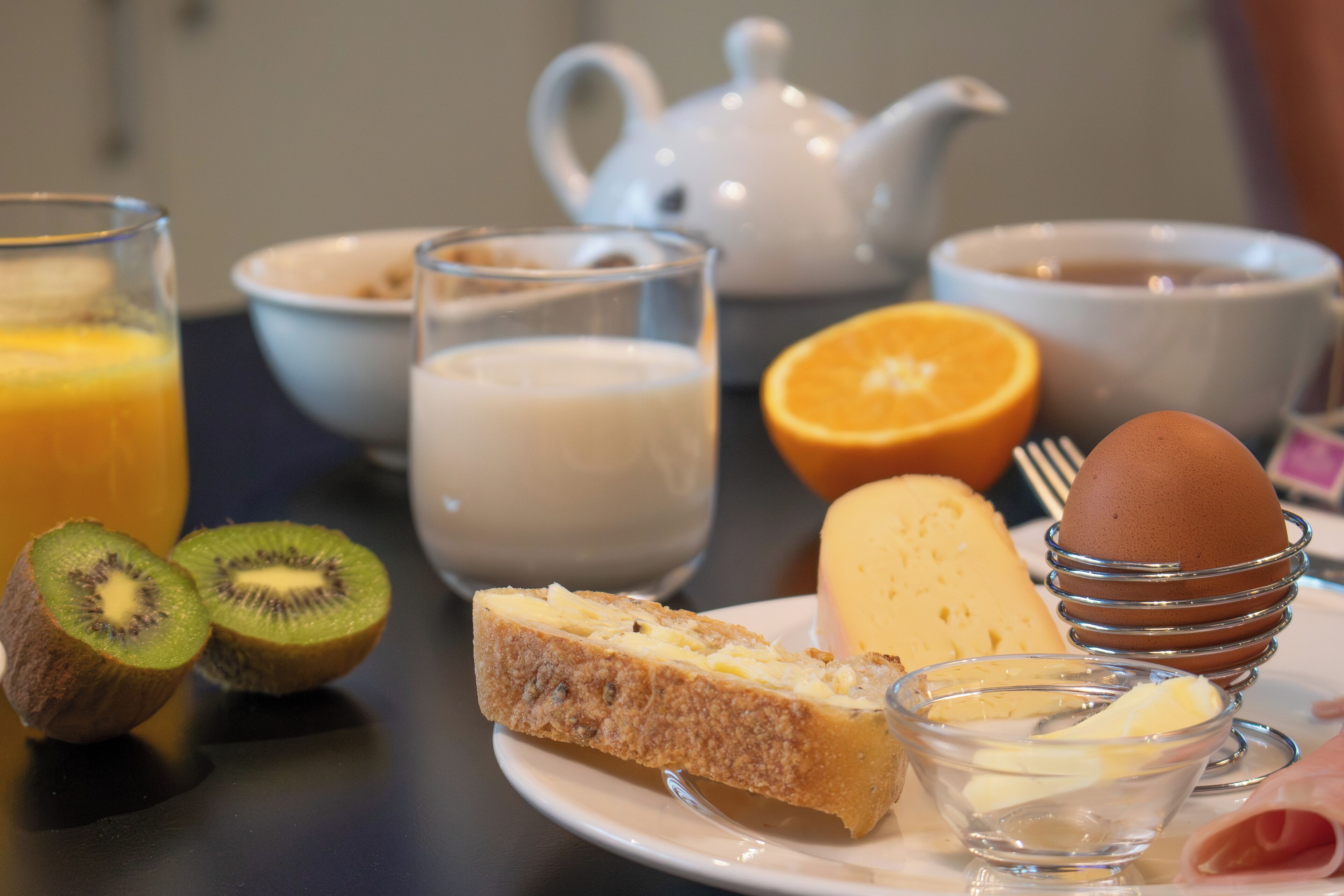 Een ontbijttafel met een theepot, kopjes, een bord met toast, een kiwi, een sinaasappel, een kommetje boter en een gekookt ei.