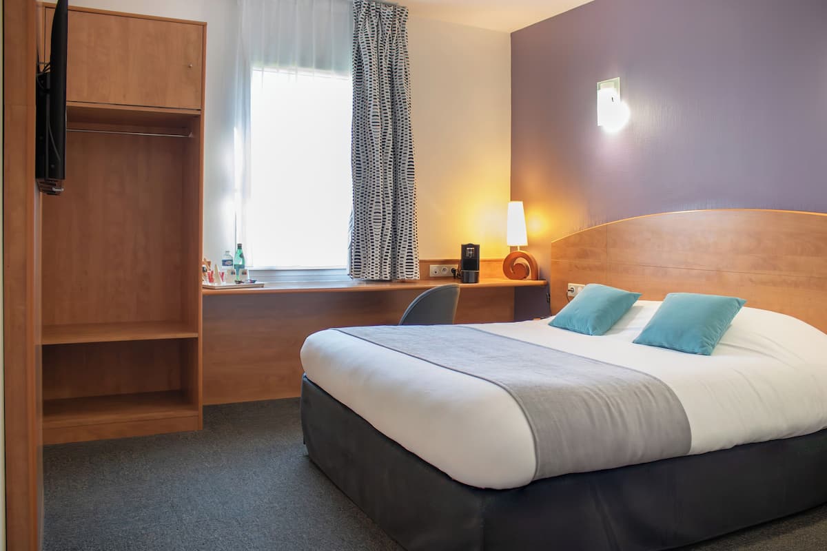 Habitación de hotel con una cama grande, un escritorio con silla, un armario y una ventana con cortinas.