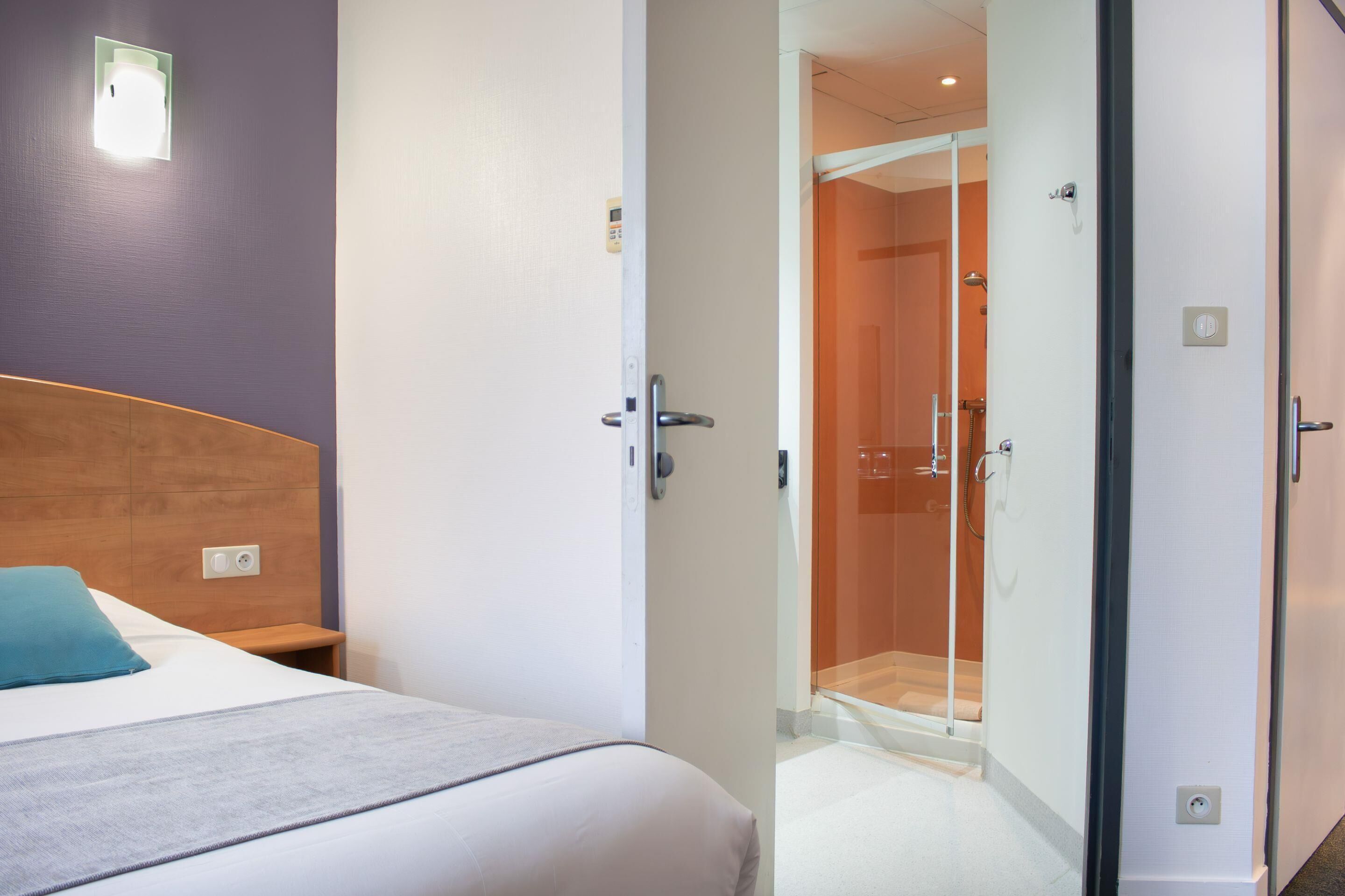 Une chambre d’hôtel comprenant un lit, une table de chevet, une salle de bain avec une douche à parois de verre et une porte donnant sur une autre pièce.