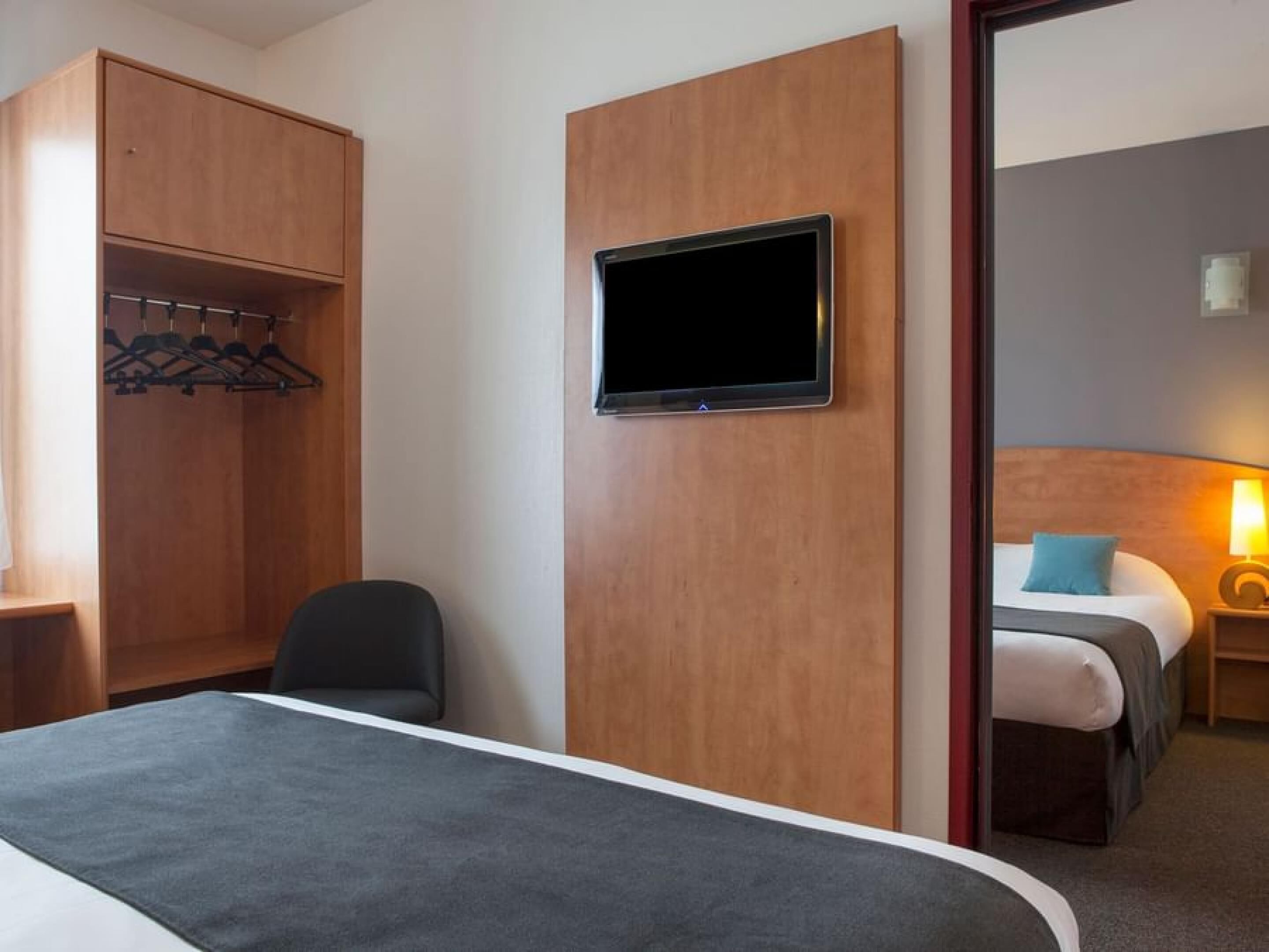 Una camera d'albergo con un letto, una TV montata su un pannello di legno, un angolo salotto con una poltrona e un armadio con abiti appesi.