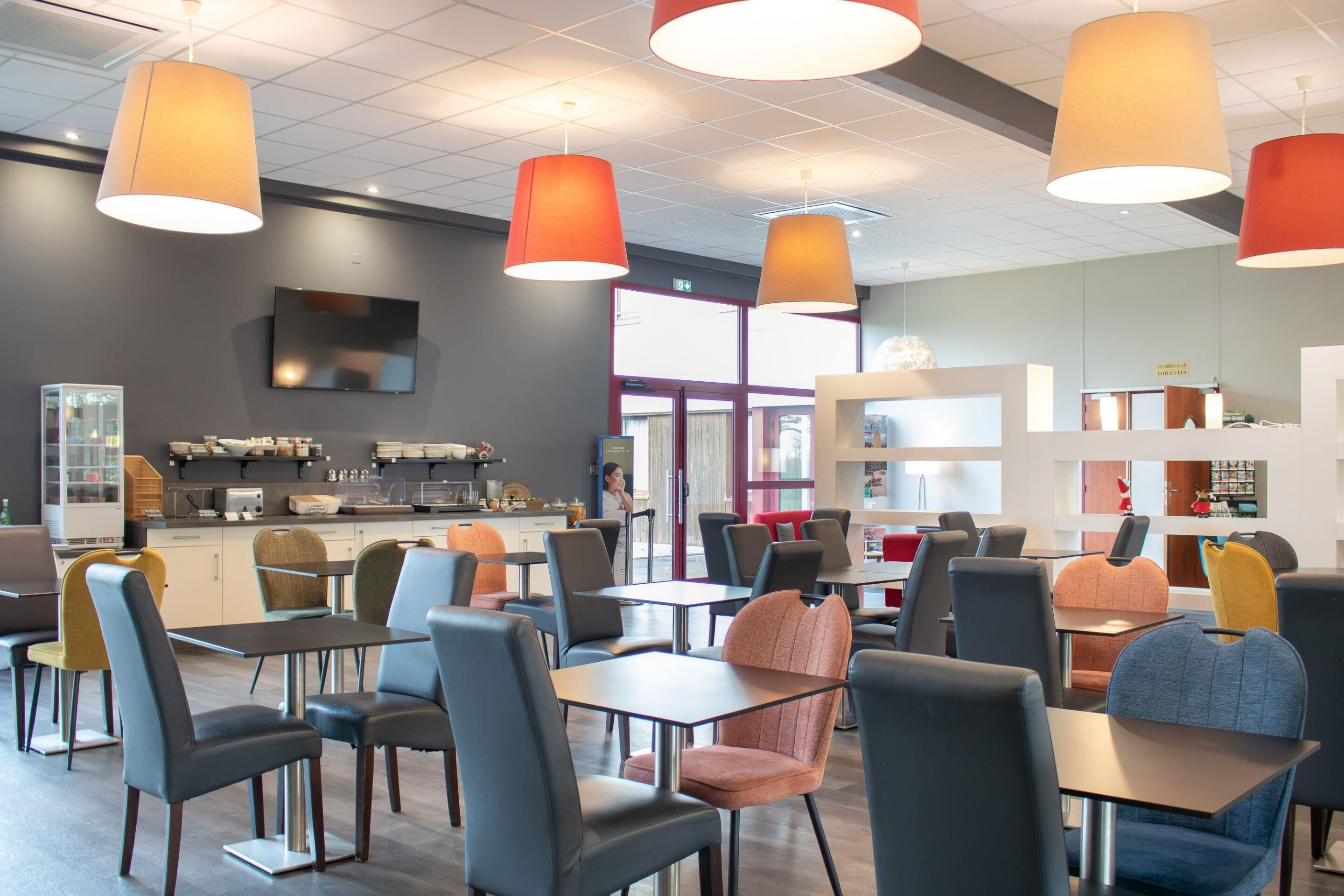 A modern cafe with multiple tables and chairs, a kitchen area, and a large window providing natural light.
