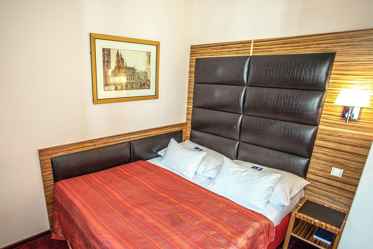 Habitación de hotel con una cama grande, cabecera de madera con paneles acolchados negros, mesita de noche pequeña, lámpara y un cuadro en la pared.