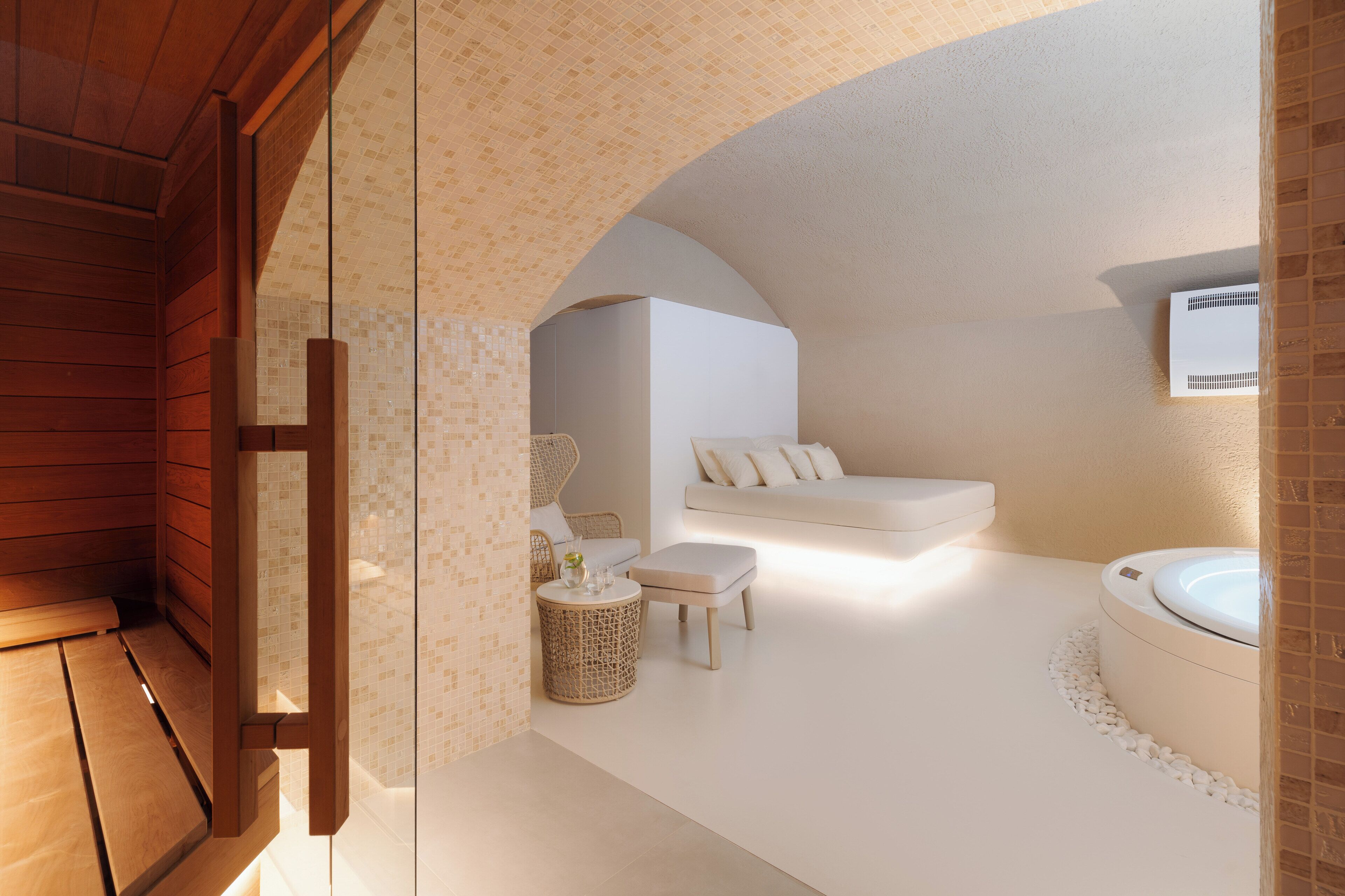 A modern spa room with a wooden bench, a wicker side table, a white sofa, and a round bathtub.