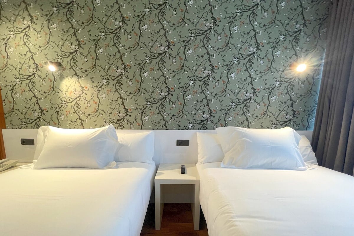Habitación de hotel con dos camas individuales, una mesita de noche, un teléfono y una pared con diseño floral.