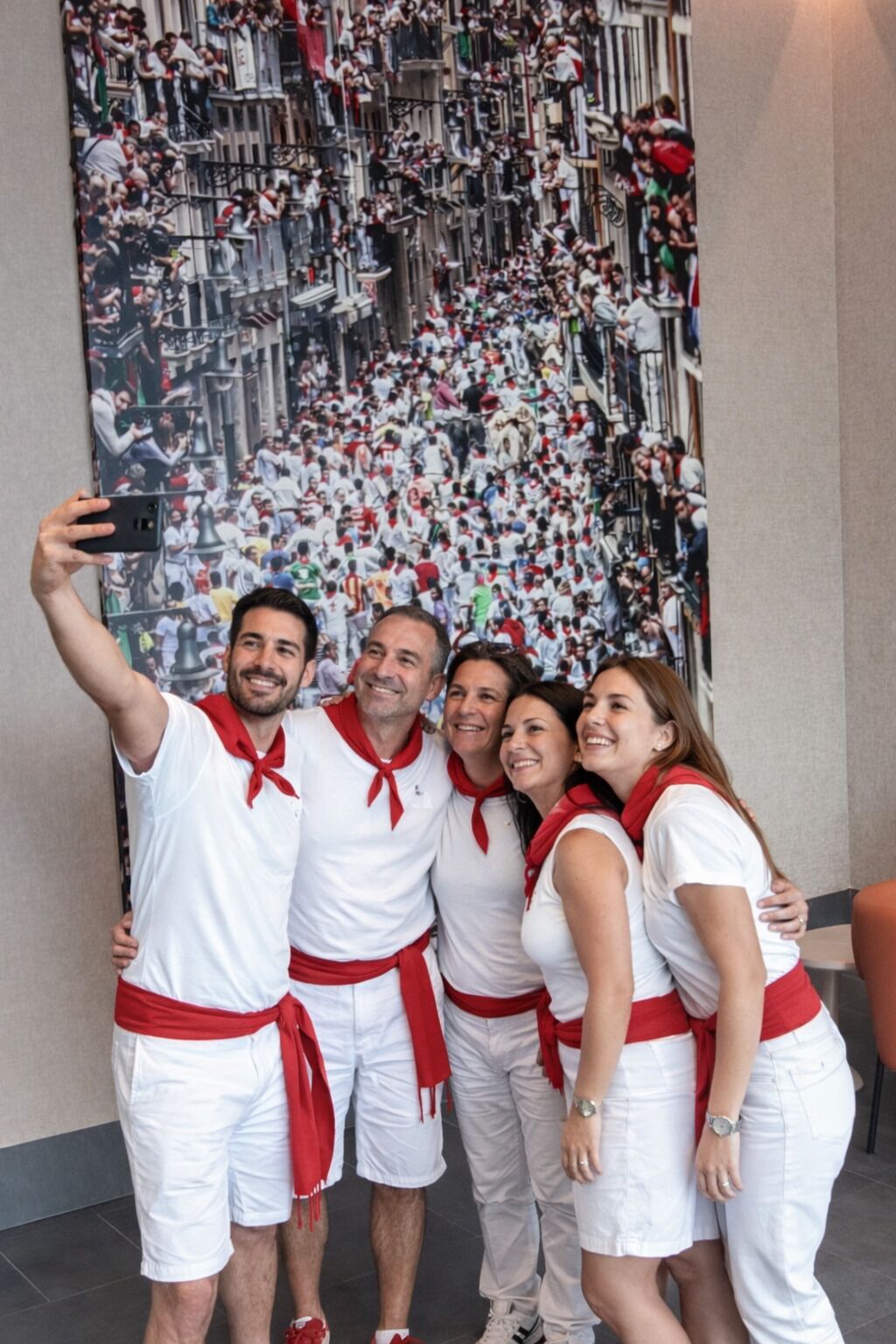 Un grupo de personas vestidas de blanco y rojo posan frente a un mural grande que muestra una escena concurrida.