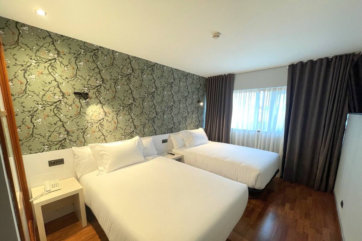 Habitación de hotel con dos camas individuales, una mesita de noche con teléfono, una pared con estampado floral y una ventana con cortinas.
