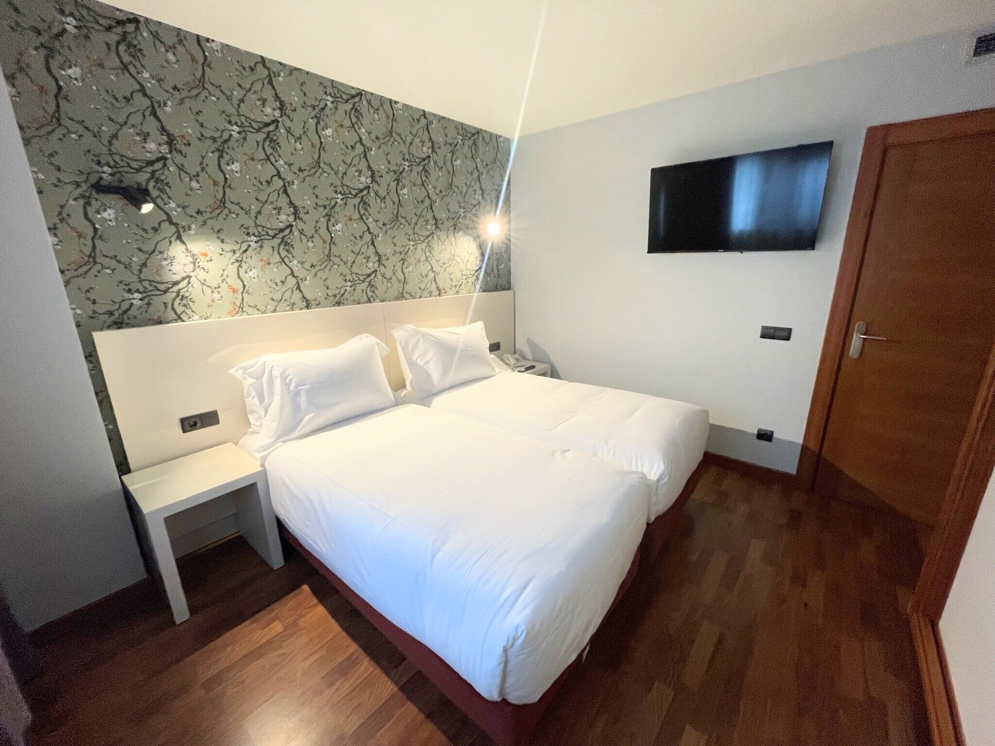 Quarto de hotel com uma cama grande, mesa de cabeceira, televisão presa na parede e chão de madeira.