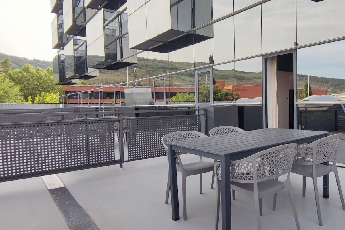 Una terraza moderna al aire libre con una mesa y sillas, y amplios ventanales que ofrecen vistas del paisaje circundante.