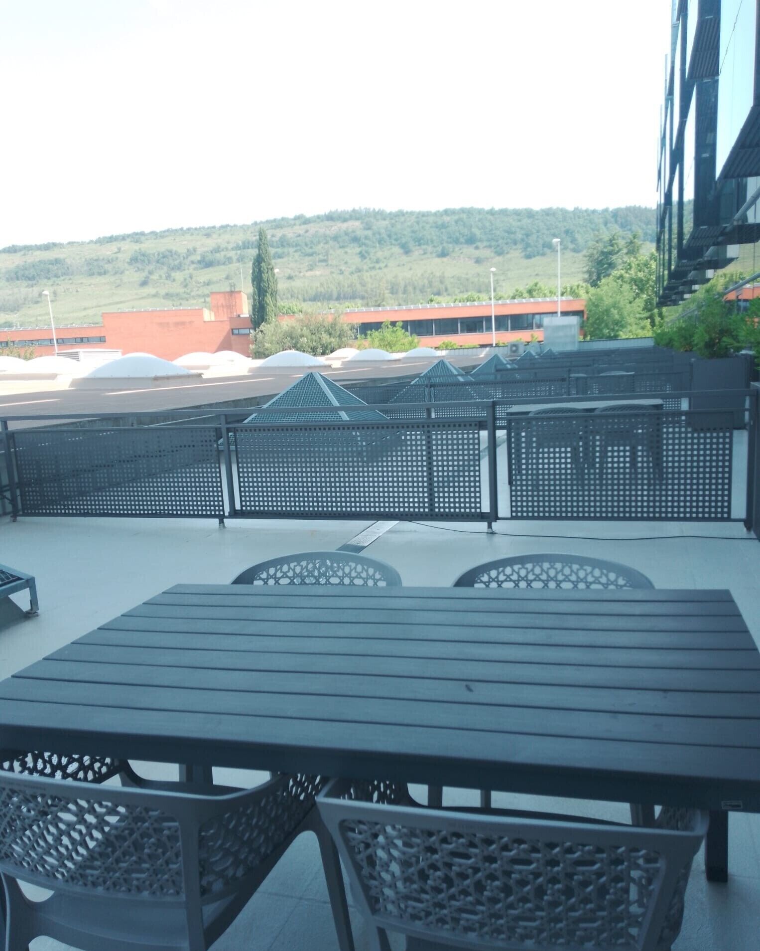 Una terraza en la azotea con una mesa y sillas, vista a un edificio de techo rojo y una ladera verde al fondo.