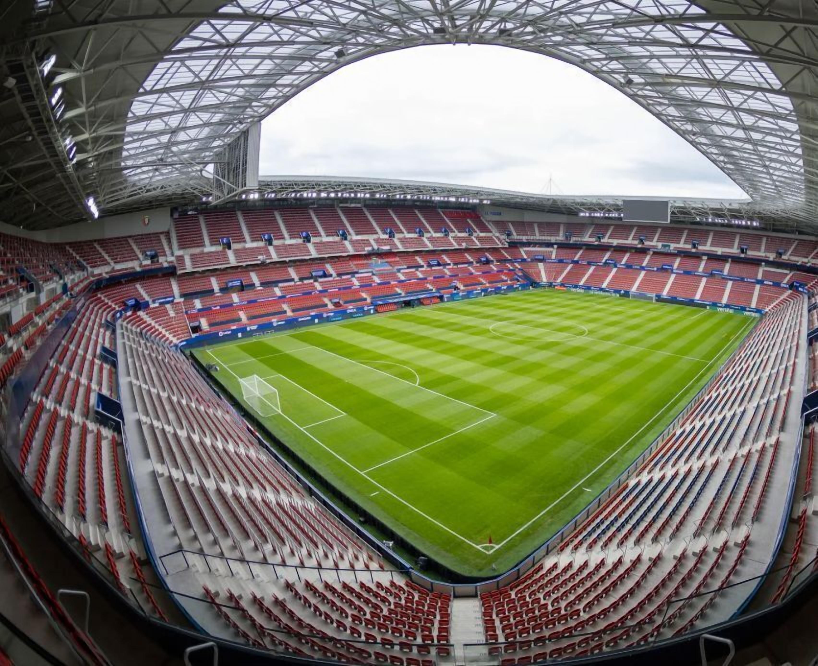 A large stadium with a green field, red and white seating, and a distinctive arched roof structure.
