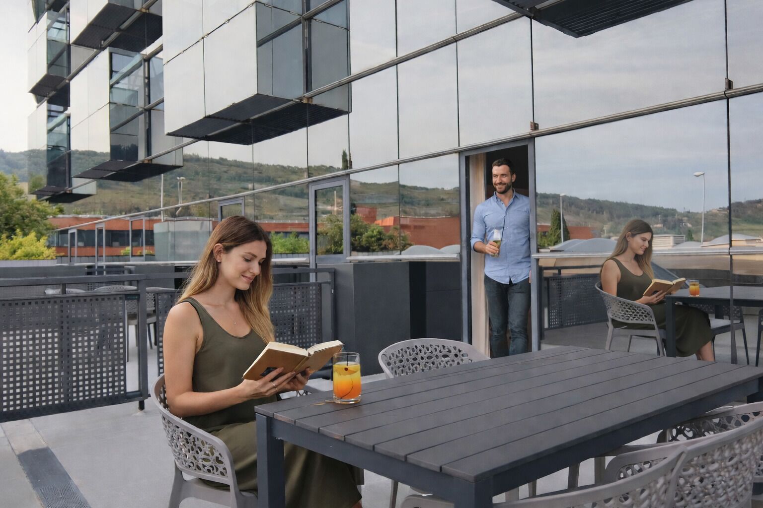 A modern outdoor seating area with a table and chairs, a glass barrier, and a view of a building with large windows.