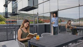 A modern outdoor seating area with a table and chairs, a glass barrier, and a view of a building with large windows.