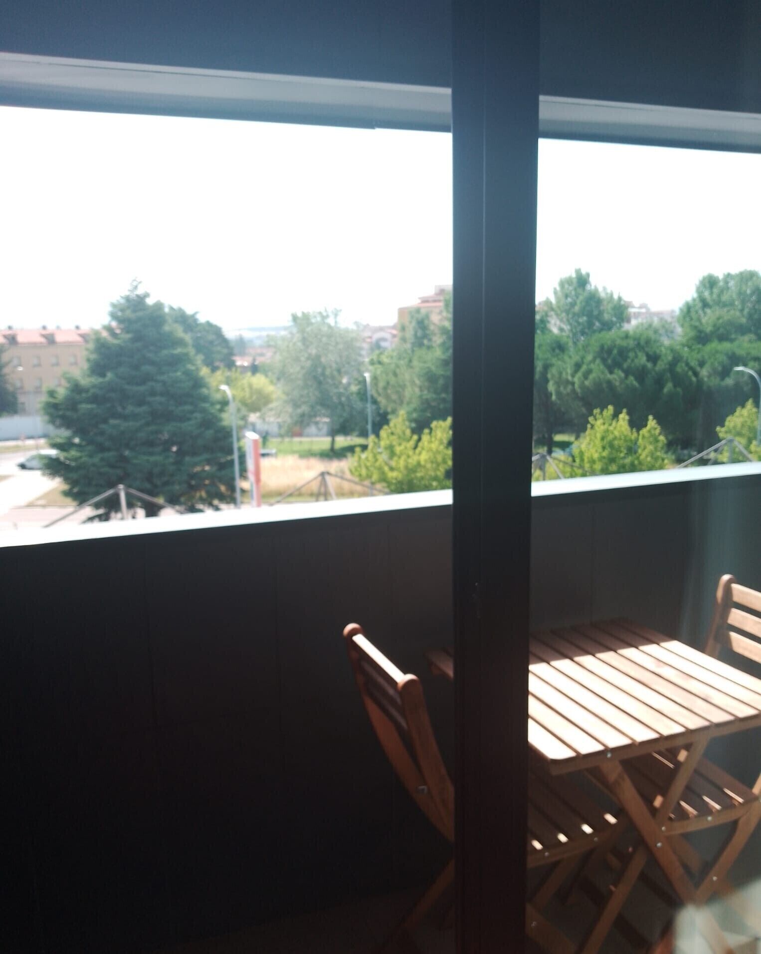 Un balcón con una mesa y sillas de madera, que da a una vista urbana con árboles y edificios.