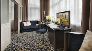 A hotel room with a dark blue sofa, a desk with a computer, a chair, and a large window with curtains. - EA Hotel Royal Esprit (Prague)