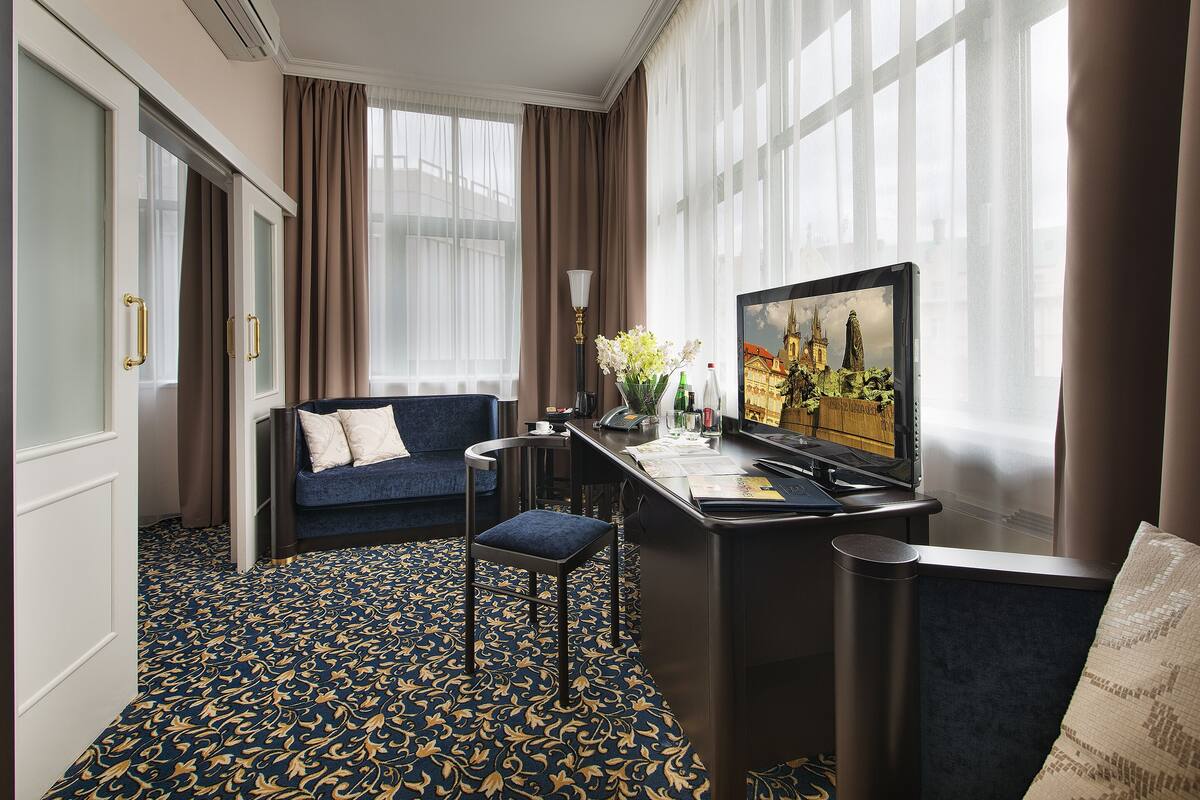Habitación de hotel con un sofá azul oscuro, un escritorio con computadora, una silla y un ventanal con cortinas.