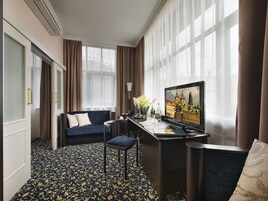 Et hotelværelse med en mørkeblå sofa, et skrivebord med computer, en stol og et stort vindue med gardiner.