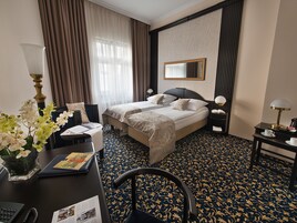 Et hotelværelse med to senge, et skrivebord med lampe, en stol, et lille bord med en kop og en dekorativ vase med blomster.