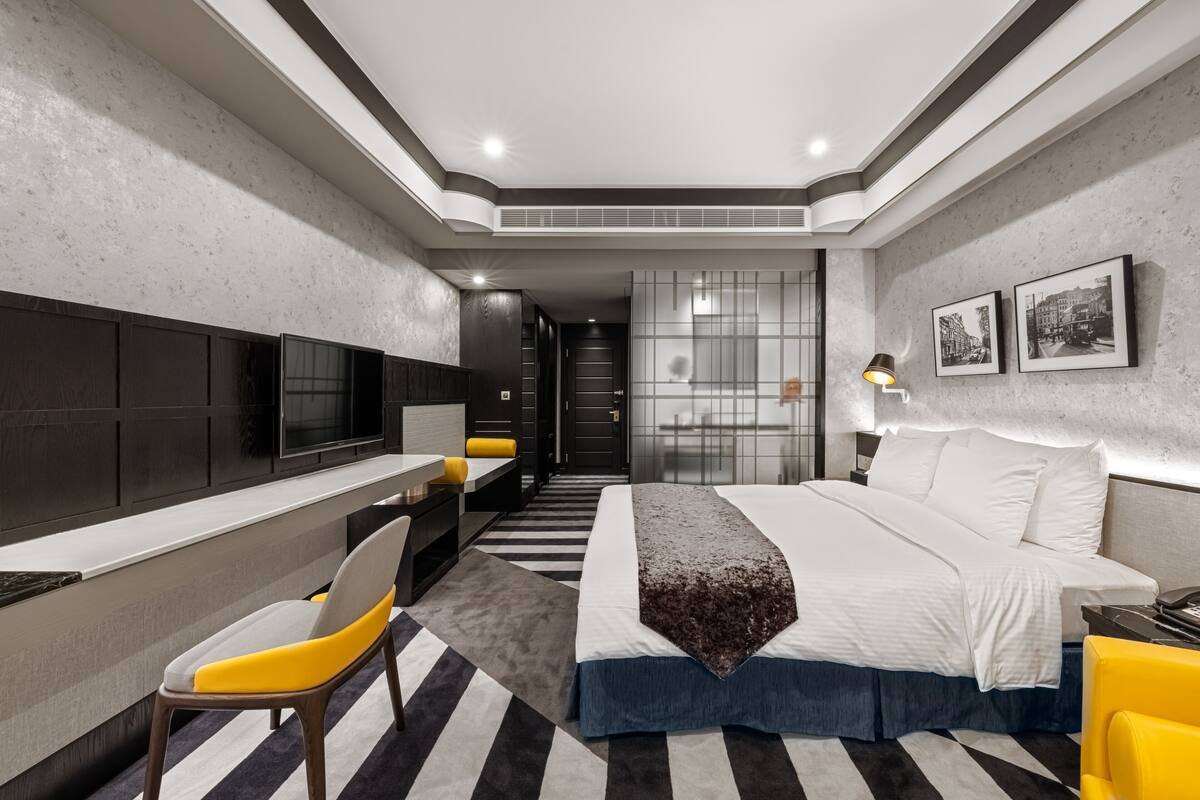 A modern hotel room with a bed, a desk, a chair, a TV, and a lamp.