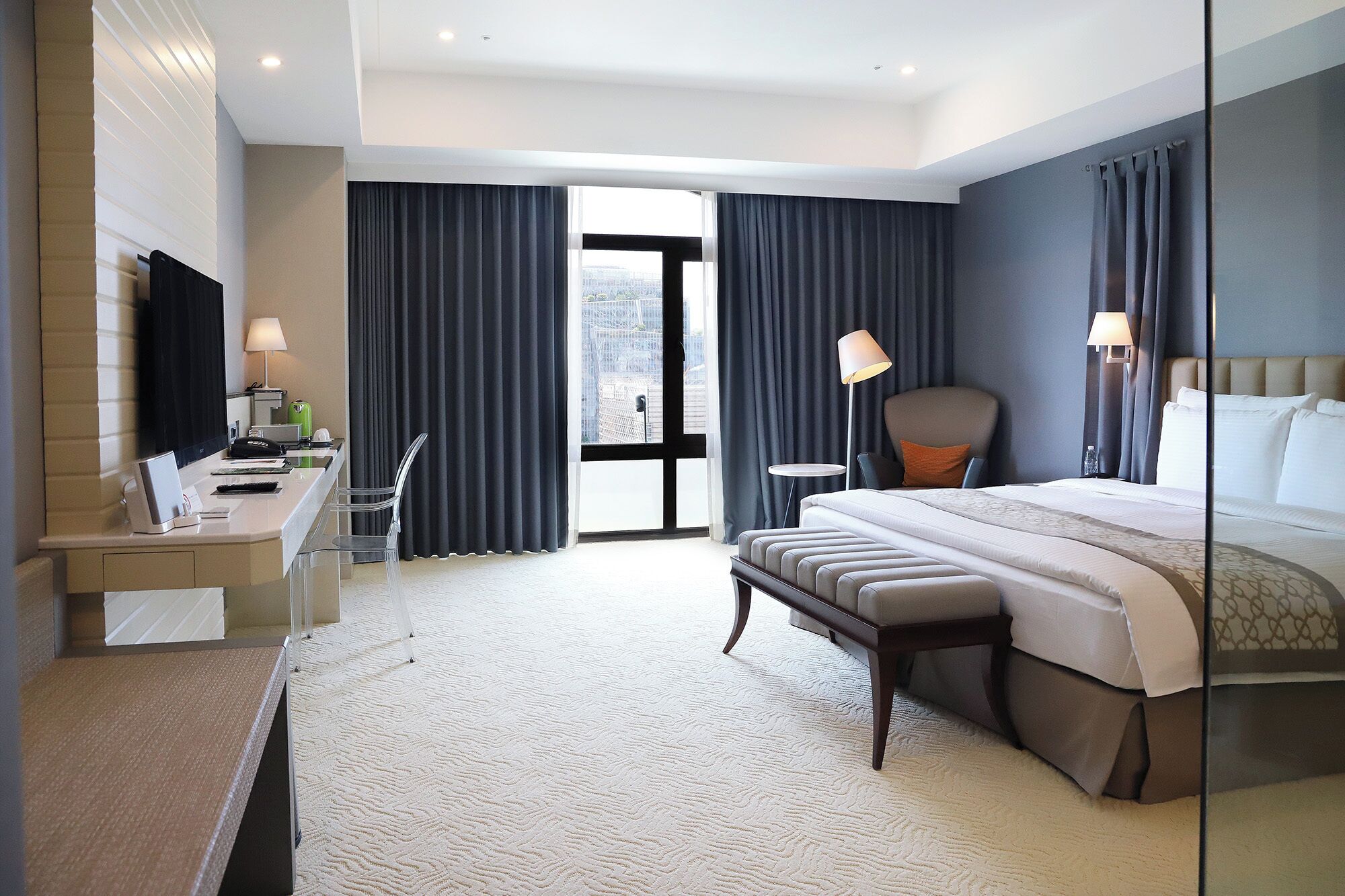A modern hotel room with a bed, bedside table, lamp, chair, and a desk with a computer.