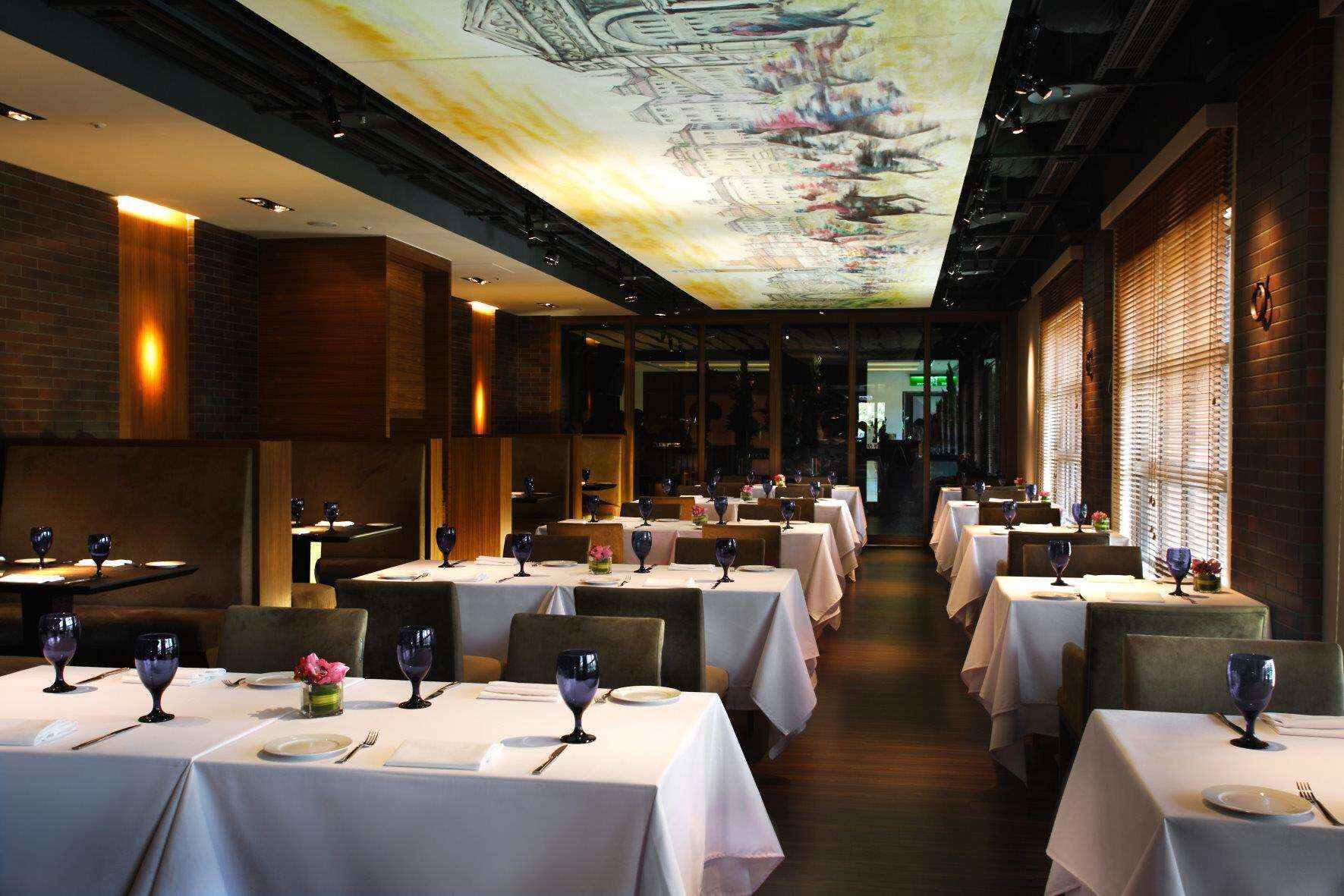 A restaurant with multiple tables set for dining, featuring a ceiling mural and brick walls.