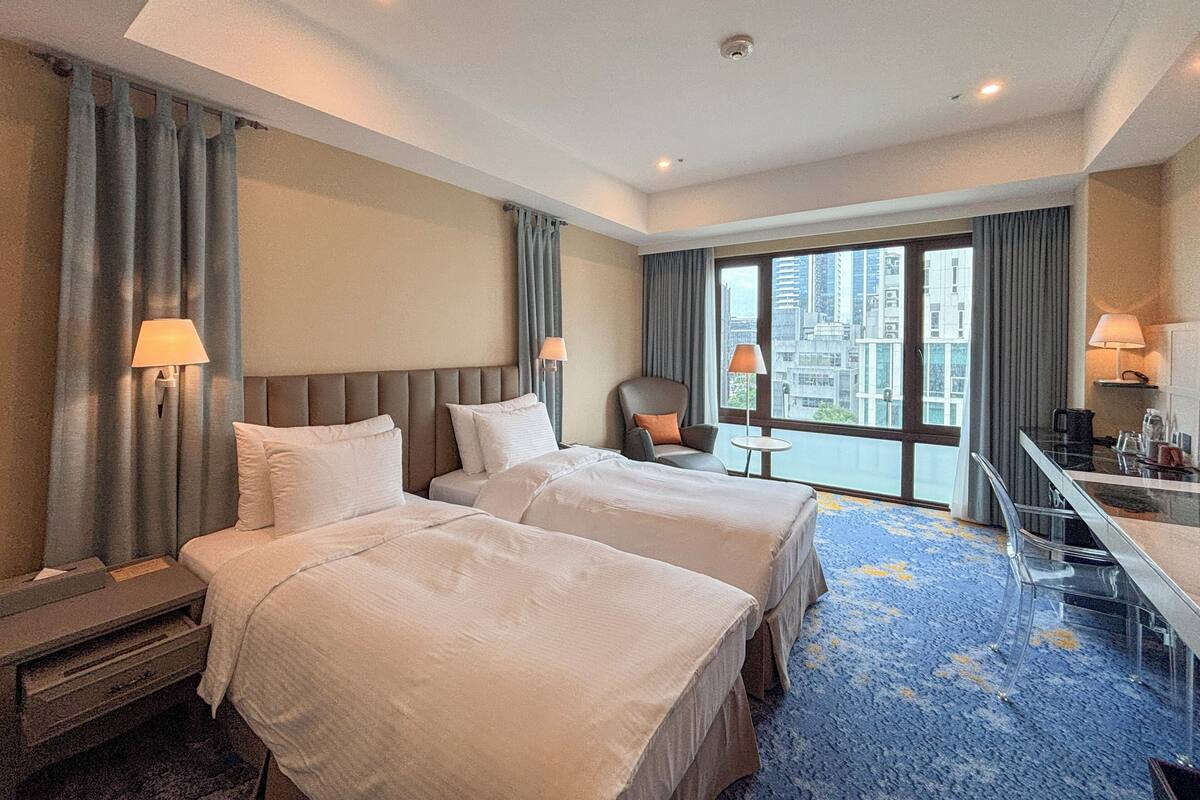 A hotel room with two beds, a desk, a chair, and a view of the city.