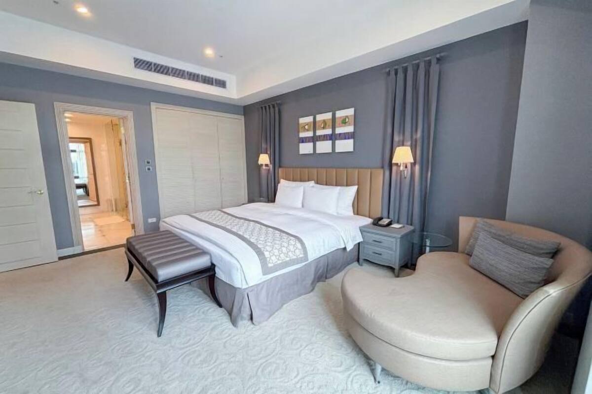 A bedroom with a bed, bedside tables, a bench, a chair, and a door leading to a bathroom.