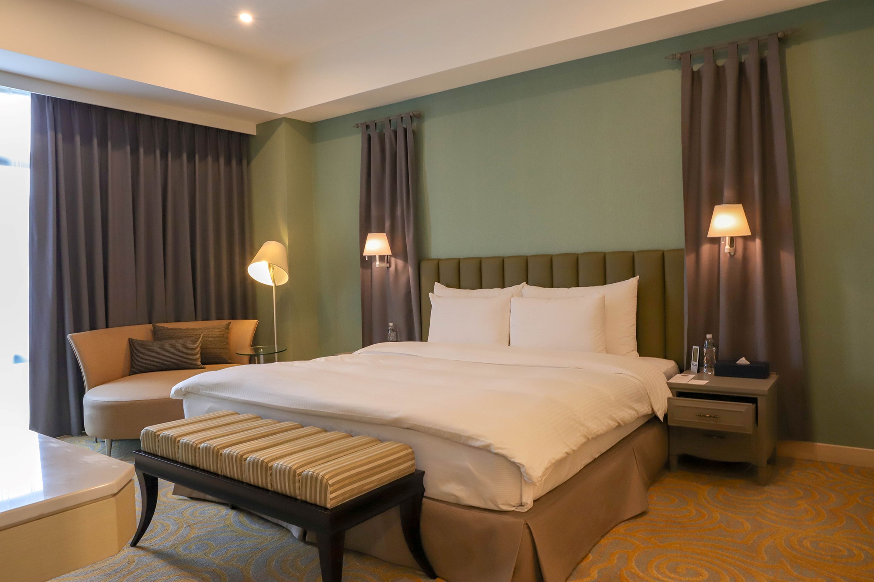 A hotel room with a large bed, a bedside table, a chair, and a window with curtains.