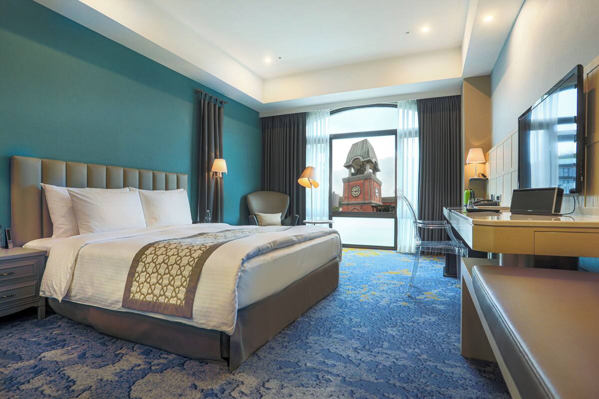 A hotel room with a large bed, a desk with a laptop, a chair, a clock tower view, and a balcony.