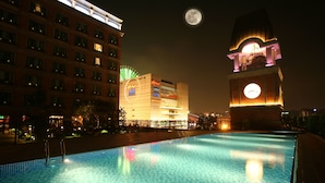 Un área de piscina nocturna con una torre del reloj y edificios iluminados de hotel al fondo.