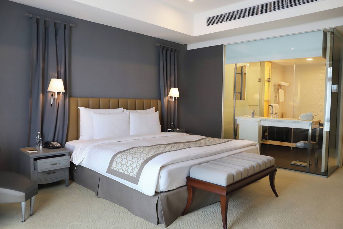 A hotel room with a large bed, bedside tables, a desk, and a bathroom visible through a glass partition.