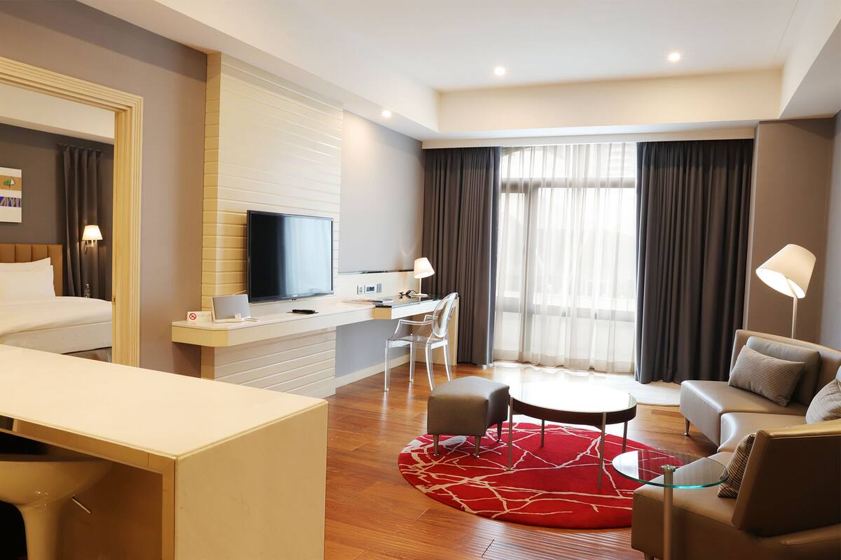 A modern hotel room with a bed, a desk with a computer, a TV, a sofa, and a dining area.