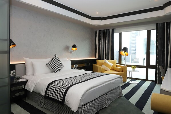 A modern hotel room with a large bed, a sofa, a small table with a plant, and a view of the city through large windows. - Grand Victoria Hotel (Taipei)