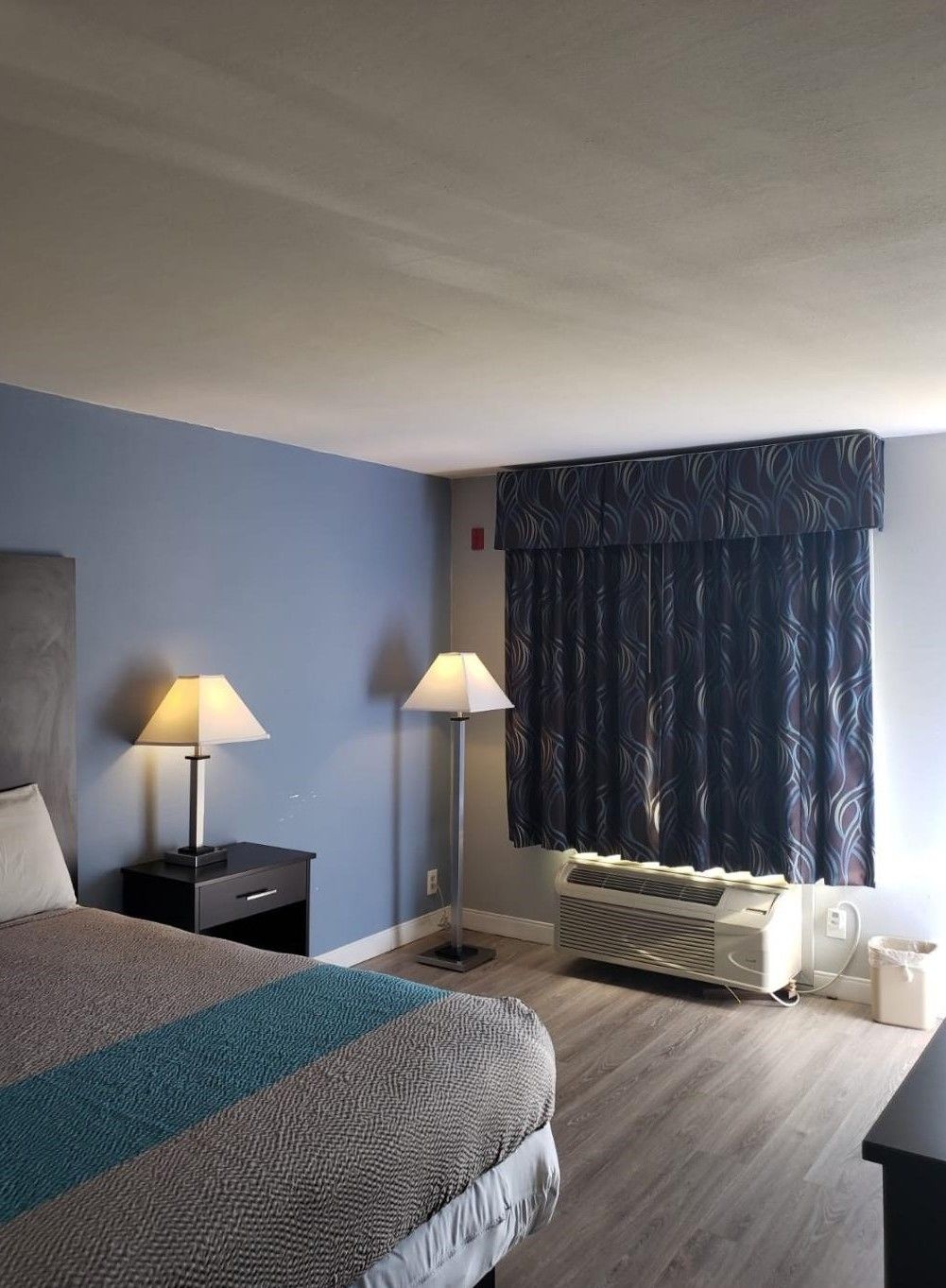 A hotel room with a bed, two bedside tables with lamps, a window with patterned curtains, and a wall-mounted air conditioner.