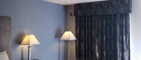 A hotel room with a bed, two bedside tables with lamps, a window with patterned curtains, and a wall-mounted air conditioner.
