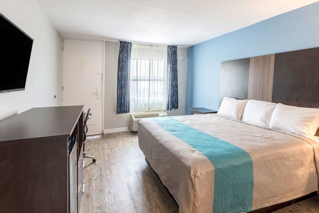 Habitación de hotel con cama, escritorio, televisor, ventana con cortinas y puerta.