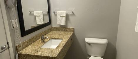Baño con lavamanos de encimera de granito, inodoro blanco, espejo y secador de pelo en la pared.