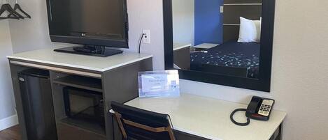 A hotel room with a desk, a computer, a telephone, and a mirror reflecting a bed.