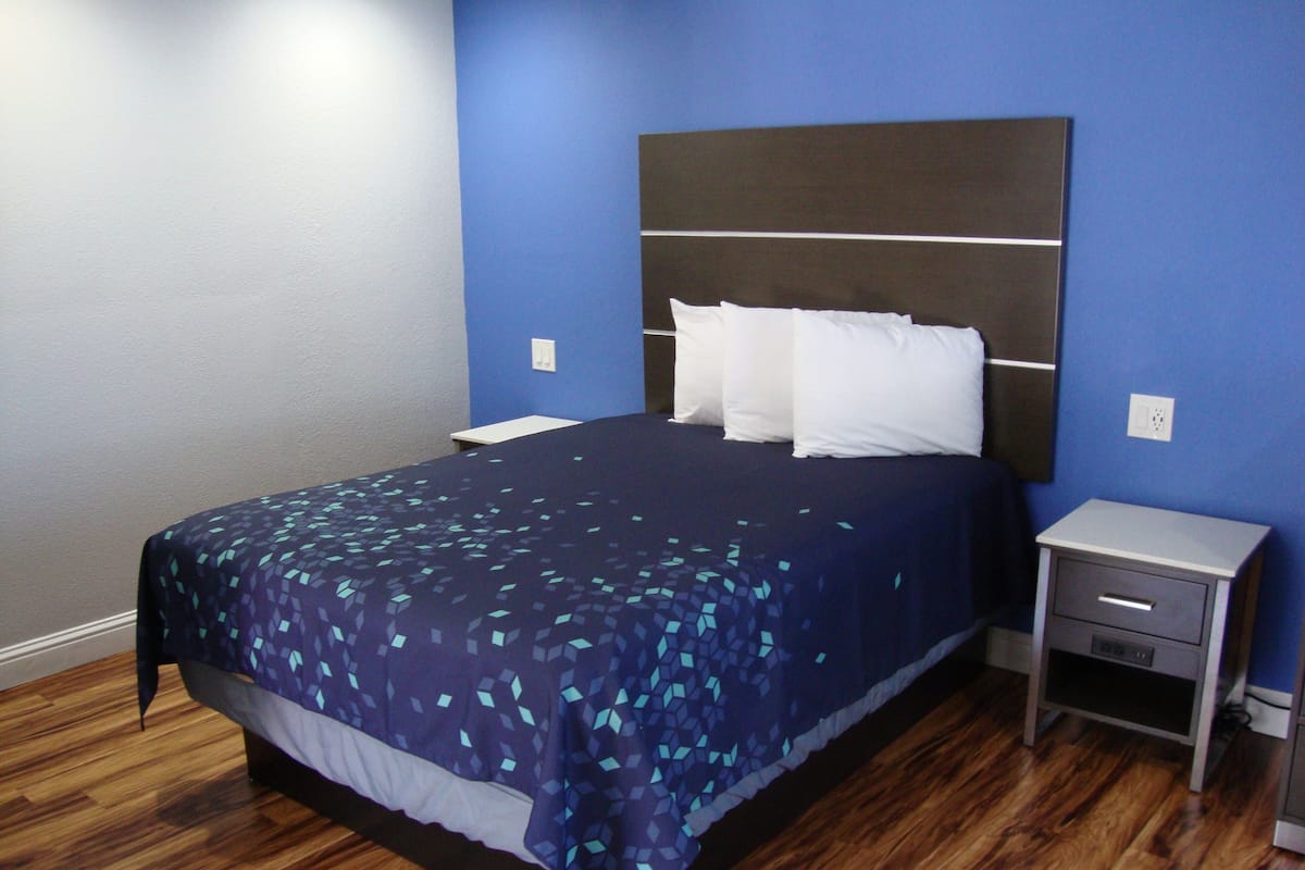 A neatly made bed with a dark blue bedspread and white pillows, a wooden floor, and a small bedside table with a drawer.