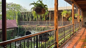 Un balcon fermé offrant une vue sur une cour intérieure, agrémentée d’une fontaine, de plantes suspendues et d’un mur en pierre.