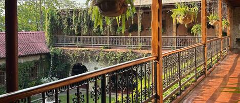 A covered balcony with a view of a courtyard, featuring a fountain, hanging plants, and a stone wall.