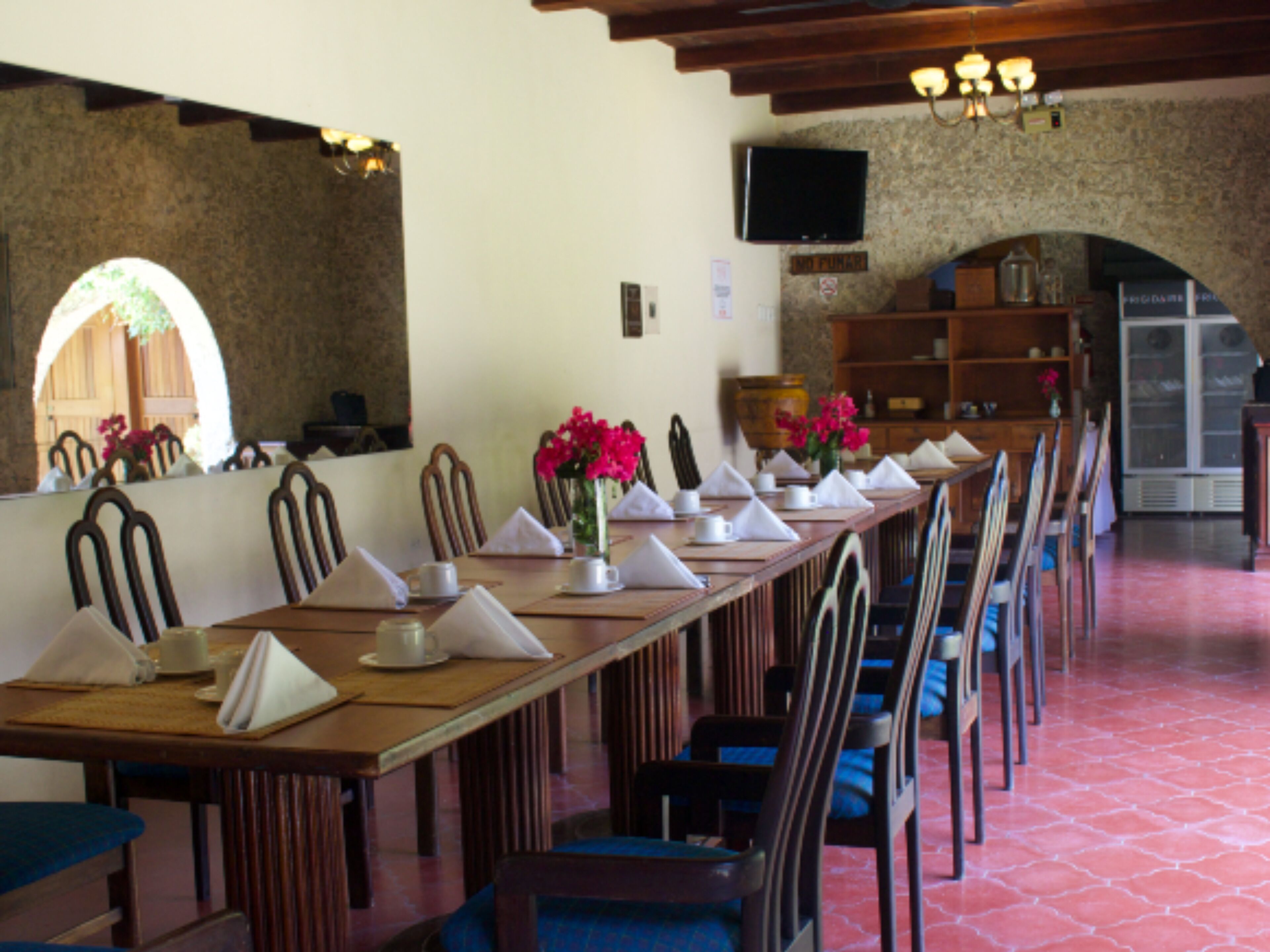 A long dining table set with chairs, a television mounted on a stone wall, and a wooden cabinet in the background.